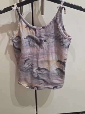 Linnetopp med delfinmotiv från Molo - Säljer en linnetopp från Molo i storlek 116 med ett coolt delfinmotiv över hela toppen. Färgerna är en mix av ljusgrå, beige och lila toner, och toppen har smala axelband med knyt. Perfekt för sommardagar och strandhäng!