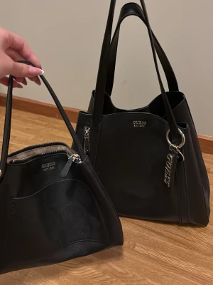Svart axelväska från Guess - Säljer en stilren svart axelväska från Guess med matchande mindre väska. Båda har silverfärgade detaljer och Guess-logga framtill. Väskorna är tillverkade i skinnimitation och har praktiska handtag samt dragkedja på den mindre väskan.