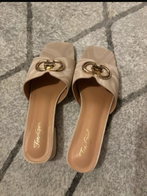 Beige sandaler med gulddetalj Ten Girl - Säljer ett par snygga beige sandaler från Ten Girl med fyrkantig tå och låg klack. Skorna har en guldfärgad metallspänne framtill och är gjorda i syntetmaterial med en mjuk innersula. Perfekt för dig som gillar stilrena och trendiga detaljer.