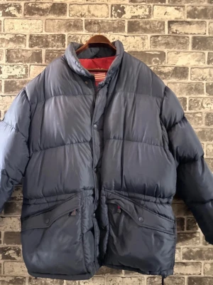 Blå pufferjacka från GANT USA Sport - Säljer en mörkblå pufferjacka från GANT USA Sport med röd insida och broderad logga på kragen. Jackan har hög krage, stora fickor med knappstängning och dragsko nedtill. Perfekt för kalla dagar och riktigt snygg streetstil.