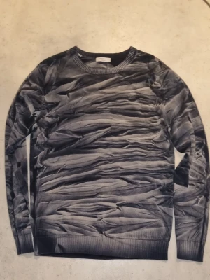 Grå stickad tröja med vågigt mönster - Säljer en grå stickad tröja med unikt vågigt mönster som ger en cool 3D-effekt. Tröjan har rund halsringning, ribbade muddar och är långärmad. Perfekt för dig som vill sticka ut med en annorlunda stil.