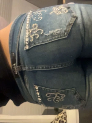 Blå jeans med strass och slitningar - Säljer ett par blå jeans från YMI med snygga strassdetaljer på fickorna och coola slitningar framtill. Jeansen har broderade mönster på bakfickorna och är i klassisk femficksmodell. Passformen är slim med lätt bootcut och materialet är stretchig denim.