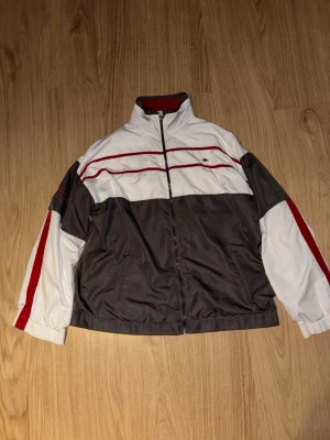 Lacoste vindjacka i vitt och grått - Snygg vindjacka från Lacoste med vit och mörkgrå färgblock, röda detaljer och broderad logga på ärmen. Jackan har hel dragkedja, hög krage och två sidofickor. Materialet är polyester och insidan har röd mesh. Perfekt för dig som gillar sportig stil.