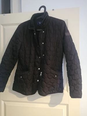 Svart quiltad jacka från GANT - Quiltad svart jacka från GANT i storlek M. Jackan har lång ärm, ståkrage och både dragkedja och knappar framtill. Snygga fickor med knappdetaljer och ett mönstrat tyg med hexagonliknande quiltning. Perfekt för dig som gillar stilren och klassisk look.