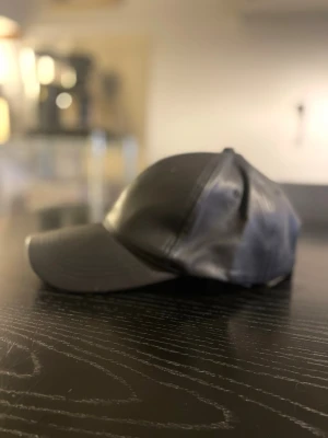 Svart designer keps från Stand Studio  - Säljer en stilren design svart keps i skinnimitation. Kepsen har klassisk form med böjd skärm och synliga sömmar för en clean look. Perfekt accessoar för att lyfta din outfit.