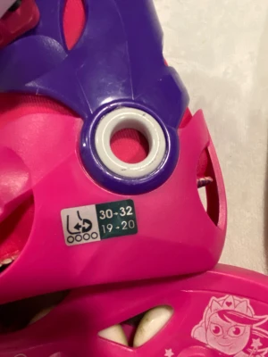 Oxelo Play3 rosa/lila rullskridskor - Säljer ett par färgglada rullskridskor från Oxelo Play3 i rosa och lila med vita hjul och coola spännen. Skorna har justerbar storlek och är dekorerade med stjärnor och en tecknad figur på sidan. Perfekt för dig som vill rulla fram med stil!