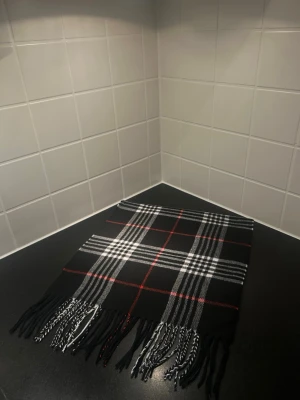 Svart rutig halsduk med fransar - Snygg svart halsduk med klassiskt rutigt mönster i vitt och rött. Halsduken har långa fransar i ändarna och är perfekt att slänga över axlarna för en cool look. Materialet känns mjukt och skönt mot huden.