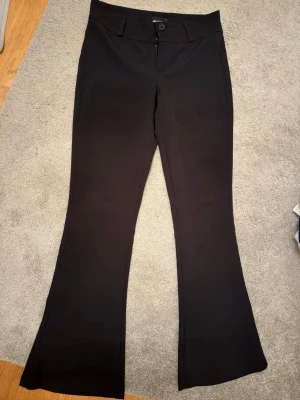 Svarta bootcut-byxor från Gina Tricot - Svarta bootcut-byxor från Gina Tricot med klassisk knappstängning och bälteshällor. Baktill finns två fickor med lock och knapp. Byxorna har en snygg utsvängd passform och är tillverkade i ett mjukt, stretchigt material som ger en bekväm känsla. Köpta för 500 och säljs för 300. Ändast använt 3 gånger. Storlek L