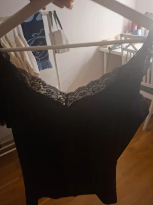 Svart linnetopp med spets - Säljer en svart linnetopp med v-ringad hals och breda axelband. Toppen har fina spetsdetaljer längs halsringningen och axelbanden som ger en extra touch. Materialet känns mjukt och följsamt, perfekt att styla med jeans eller kjol.