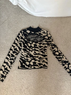 Svart och beige stickad tröja med mesh - Säljer en cool långärmad stickad tröja med svart och beige zebramönster. Tröjan har en hög krage och en unik meshdetalj över bröstet som ger en edgy vibe. Perfekt för dig som vill sticka ut med din stil.