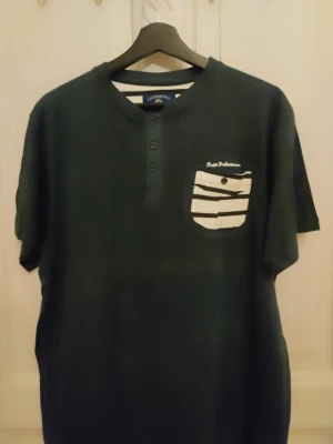 Svart Performance t-shirt med randig ficka - Helt ny svart t-shirt från Performance, modell RCMN Crew, med korta ärmar och rund hals. Framsidan har en vit och svart randig bröstficka med knapp och tryckt logga ovanför. T-shirten har tre knappar vid halsen och är gjord i mjuk bomull. Perfekt för chill och streetwear.