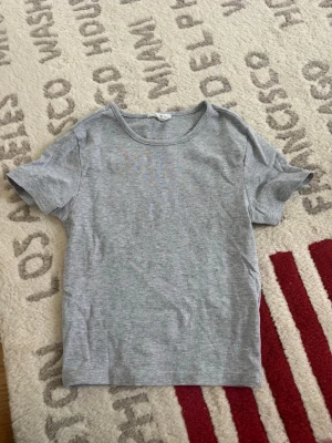 Grå basic t-shirt från Gina Tricot  - En enkel grå t-shirt från Gina Tricot i mjuk bomull. Modellen är klassisk med rund hals och korta ärmar, perfekt för en chill och clean look. Passformen är normal och tyget är tunt och skönt mot huden.