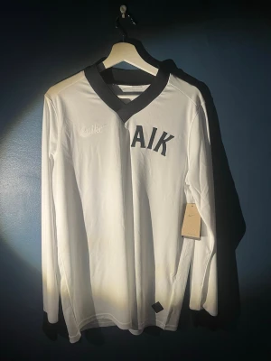 AIK 1924 Edition Målvaktströja  - Snygg AIK fotbollströja från Nike i vit Dri-Fit polyester med svart V-ringad krage och muddar. Tröjan har broderad Nike-logga och AIK-tryck på bröstet, samt texten 'Allmänna Idrottsklubben' i nacken. Slim fit-modell som passar perfekt för match eller träning. Original Pris: 1299kr Skriv om ni har några funderingar kring tröjan
