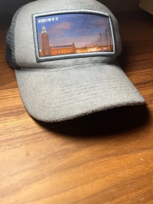 Grå keps från Revenite Pop Up '23 - Säljer en grå keps från Revenite Pop Up '23 med en snygg bild på Stockholms stadshus framtill. Kepsen har mesh på baksidan och insidan är dekorerad med Revenite-tryck. Materialet är mjukt och har en lite ruggad yta. Knappt använd och kommer i riktigt bra skikt.