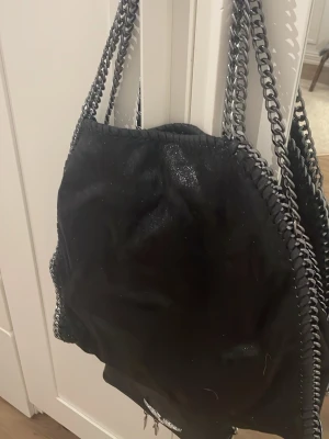 Svart axelväska med kedjedetaljer - Säljer en svart axelväska med coola silverfärgade kedjor som axelrem och runt kanterna. Väskan har en mjuk, glansig yta som ger en snygg look och känns lyxig. Perfekt för dig som gillar att sticka ut med detaljer och vill ha en väska som rymmer allt du behöver.