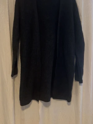 Svart stickad kofta med lång ärm - Mysig svart stickad kofta med lång ärm och öppen framtill. Perfekt att slänga över en t-shirt eller topp när det är lite kyligt. Koftan har en loose passform och är tillverkad i ett mjukt stickat material som känns skönt mot huden.