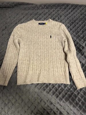 ÄKTA Grå stickad Ralph lauren tröja - Säljer en ÄKTA grå stickad tröja från Polo Ralph Lauren med klassiskt kabelstickat mönster och rund hals. Tröjan har långärmade ribbade muddar och en broderad logga på bröstet. Perfekt för dig som gillar stilrena och tidlösa plagg.