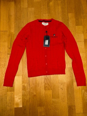 Röd kofta från Bondelid XS - Säljer en snygg röd kofta från Bondelid i storlek XS. Koftan är kabelstickad med rund hals, knappar framtill och broderad logga på bröstet. Perfekt för dig som gillar klassisk stil med lite färg. Materialet är mjukt och skönt.