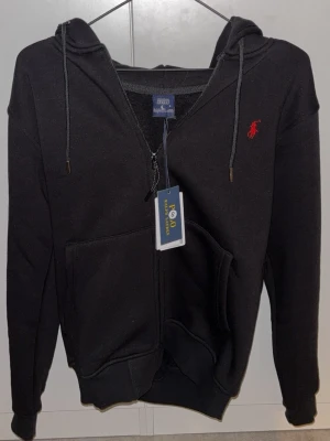 Svart hoodie från Polo Ralph Lauren och beige - Svart hoodie från Polo Ralph Lauren med röd broderad logga på bröstet. Tröjan har huva med snörning, magficka och dragkedja framtill. Tillverkad i mjuk bomull och har långa ärmar. Perfekt för chill dagar. En ligger på 899 kr men pris kan diskuteras privat!