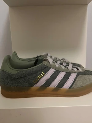 Adidas Gazelle sneakers grön/lila - Adidas Gazelle sneakers i grön mocka med ljuslila ränder och snörning. Sulan är i gummi med en brun ton och skorna är ruggat mocka. Andvända en gång.