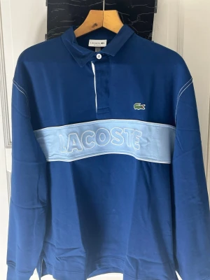 Blå pikétröja från Lacoste - Snygg blå pikétröja från Lacoste med vit knapp och klassisk krage. Tröjan har ett brett ljusblått band över bröstet med stor broderad Lacoste-logga och den ikoniska krokodilen på vänster sida. Långärmad och tillverkad i mjuk bomull.