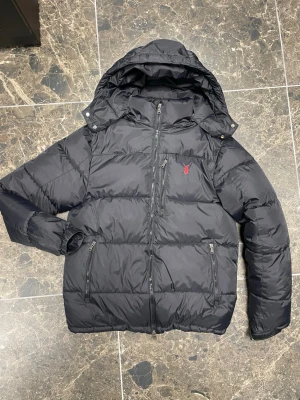 Svart pufferjacka från Polo Ralph Lauren - Svart pufferjacka från Polo Ralph Lauren med huva och dragkedja framtill. Jackan har tre zip-fickor och den klassiska röda Polo-loggan broderad på bröstet. Materialet är syntet och dun, perfekt för kalla dagar. Stilen är clean och jackan är långärmad med normal passform.