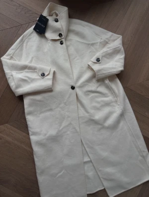 Vit Mango kappa - Jag säljer en elegant kappa från Mango som aldrig är använd med lappen kvar. Kappan är i en benvit färg och har en hög krage. Den är i ett super fint material, bestående av 50% ull. Helt slutsåld på hemsidan😊