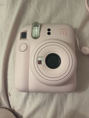 Instax Mini 12 - Fujifilm Instax Mini 12 är en kompakt och stilren direktbildskamera i ljusrosa färg. Kameran har en 60mm lins och inbyggd blixt, perfekt för att ta omedelbara foton. Skicket ser mycket bra ut utan synliga repor eller skador. Enkel att använda och idealisk för spontana minnen.