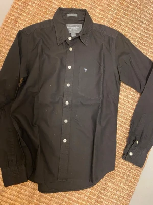 Svart skjorta Abercrombie & Fitch S - Svart skjorta från Abercrombie & Fitch i storlek S, muscle fit. Klassisk krage, vita knappar och liten bröstficka med broderad älg. Skjortan är långärmad och tillverkad i mjuk bomull som känns skön mot huden. Perfekt för dig som gillar stilrena och enkla plagg.