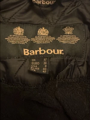 Svart quiltad jacka från Barbour - Quiltad svart jacka från Barbour med klassisk dragkedja och tryckknappar i metall. Jackan har långärmade ärmar och ett stilrent rutmönster över hela ytan. Perfekt för dig som gillar tidlös design och vill ha något som funkar till många olika looks.