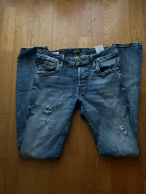 Blå slitna jeans från Jack & Jones - Säljer ett par blå jeans från Jack & Jones med snygga slitningar och hål på benen. Klassisk femficksmodell med normal passform och knappgylf. Jeansen har en cool tvättad look och är gjorda i bomullsmaterial. Perfekt för dig som gillar en avslappnad stil. Köp för 700kr. Dm mig för mer bilder eller frågor. Pris kan diskuteras
