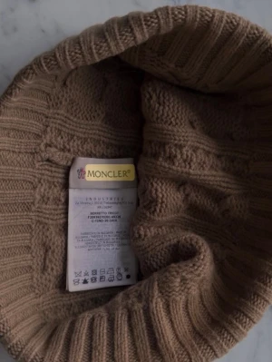 Beige Moncler mössa med pälsboll - Säljer en beige stickad mössa från Moncler med flätmönster och stor pälsboll på toppen. Mössan har en bred ribbad kant och Moncler-logga framtill. Materialet är en lyxig blandning av kashmir och ull, vilket gör den supermjuk och varm.