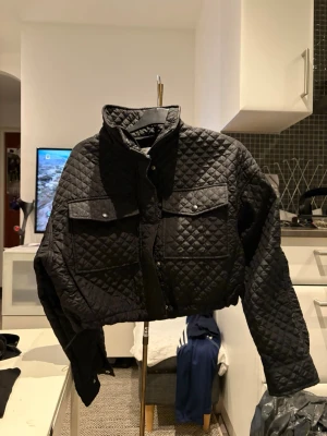 Svart quiltad bomberjacka från Bershka - Quiltad svart bomberjacka från Bershka med croppad passform, två stora fickor med lock och knappar framtill. Jackan har hög krage och dragkedja samt tryckknappar. Perfekt för dig som gillar streetstyle och vill ha en edgy look.