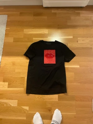 Svart t-shirt från limitato - Svart t-shirt från LIMITATO med korta ärmar och en stor röd glitterläpp på bröstet. Trycket är placerat på en glittrig röd rektangel som ger en cool och unik look. Materialet känns som mjuk bomull och passformen är normal. Perfekt för dig som vill sticka ut. Aldrig använd 