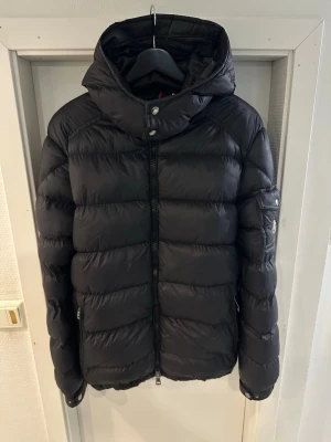 Svart Moncler maya - Säljer en svart pufferjacka från Moncler med huva och snygga breda kanaler. Använd fåtal gånger då den inte passa