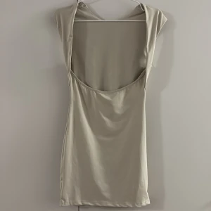 Beige kortklänning med öppen rygg - Snygg beige kortklänning med stilren design och öppen rygg. Klänningen är ärmlös och har en mjuk, slät yta som ger en modern och fräsch look. Perfekt för dig som gillar minimalistisk stil och vill sticka ut med en enkel men cool detalj.