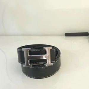 Svart skinnbälte med H-spänne - Säljer ett svart bälte med ett stort H-format metallspänne. Bältet är gjort i äkta skinn med vit kontrastsöm längs kanterna och har en snygg, klassisk design som passar till jeans eller kostymbyxor. Dma innan ni köper eller vid intresse!!