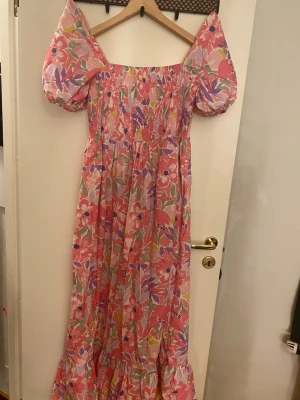 Blommig långklänning från n&u by Effdal - Säljer en färgglad långklänning med puffärmar från n&u by Effdal. Klänningen har ett blommigt mönster i rosa, lila, rött och grönt, med smock upptill och volang längst ner. Materialet känns lätt och luftigt, perfekt för varma dagar.