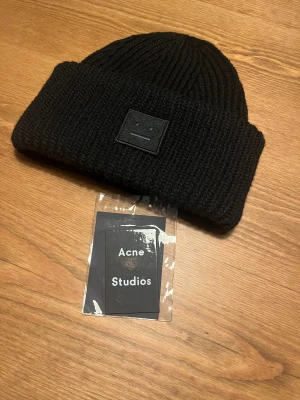 Svart stickad mössa Acne Studios - Säljer en svart stickad mössa från Acne Studios med den ikoniska ansiktsloggan framtill. Mössan har bred uppvikt kant och är tillverkad i mjukt stickat material som håller dig varm. Perfekt accessoar för kalla dagar och snygg till streetwear.
