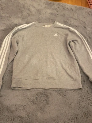 Grå sweatshirt från Adidas - Säljer en grå sweatshirt från Adidas med klassiska vita ränder längs ärmarna och Adidas-logga på bröstet. Tröjan har rund halsringning och långa ärmar. Perfekt för en sportig och avslappnad stil. Kan eventuellt tänka mig gå ner i pris