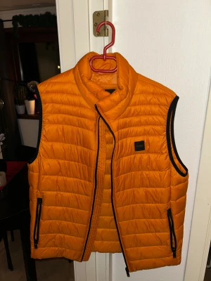 BOSS väst  - Säljer en riktigt snygg orange dunväst från BOSS med svarta detaljer och dragkedjor. Västen har quiltad design, hög krage och två praktiska fickor med dragkedja. Perfekt för dig som vill sticka ut med färg och stil under kyliga dagar.