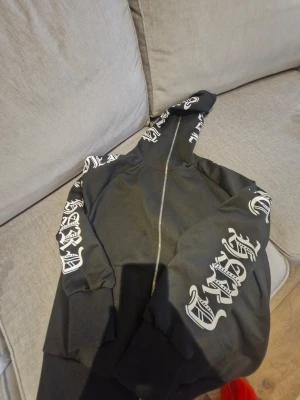 Hoodie med tryck  - Hoodie med tryck på armarna och huvan, dragkedja hela vägen upp till ansiktet, oanvänd. Storlek Xs/S