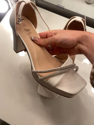 Vita sandaler med klack och strass - Snygga vita sandaler från Bocade med bred klack och fyrkantig tå. Skorna har två tunna remmar med glittrande strassdetaljer och en justerbar rem runt ankeln. Perfekta för att lyfta din outfit till nästa nivå. 🤍