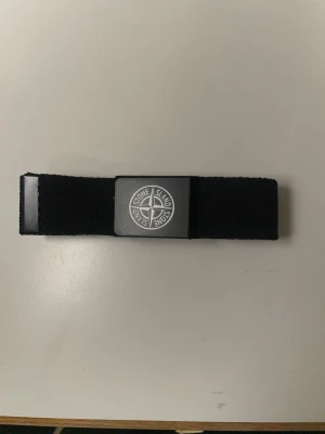 Feet Svart bälte  - Säljer ett svart bälte från Stone Island med klassisk logotyp på metallspännet. Bältet är gjort i ett slitstarkt syntetmaterial och har en enkel, stilren design som passar till de flesta outfits. Perfekt accessoar för dig som gillar streetwear.