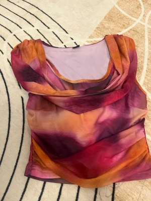 Topp - Säljer en blus i mesh med draperad front och vacker färgskiftning i lila, orange och rosa toner. Blusen har ärmlös design och ett tunt, transparent material som ger en cool och trendig look. Perfekt för dig som gillar att sticka ut med färg och stil.