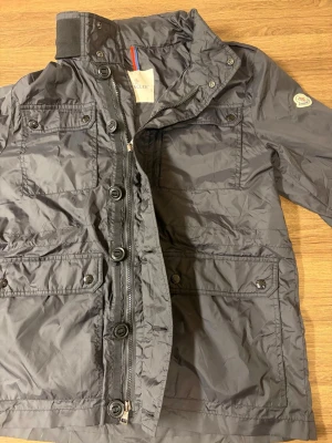  vindjacka från Moncler - Snygg grå vindjacka från Moncler med flera fickor framtill, både med knapp och dragkedja. Jackan har hög krage, Moncler-logga på ärmen och coola detaljer som stora knappar och dragkedja längs fronten. Perfekt för dig som gillar stilren och funktionell design.