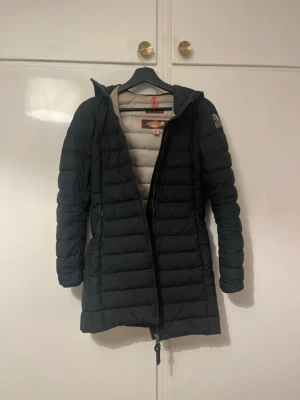 Svart dunjacka Parajumpers XS - Lång, svart dunjacka från Parajumpers i modellen Irene Woman. Jackan har quiltad design, huva och dragkedja framtill. Superlätt och varm, perfekt för kyliga dagar. Märke på ärmen och snygg passform som sitter riktigt bra. Finns fyra fickor utanpå jackan varav en dragkedja låssnat så det ej går att stänga. Jackan är även lite smutsig men går att tvätta bort!