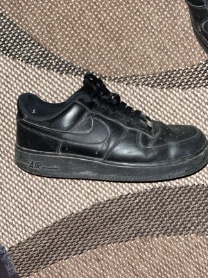 Svarta Nike Air Force 1 sneakers - Klassiska helsvarta Nike Air Force 1 sneakers i skinn med rund tå och snörning. Snygg streetstil med perforerade detaljer på tån och ikonisk Nike swoosh på sidan. Passar perfekt till jeans eller shorts för en clean och tidlös look.