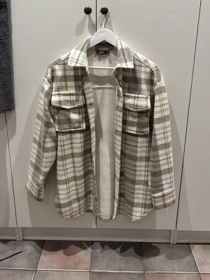 Rutig kappa  - Säljer en snygg rutig kappa från Kappahl i beige, grå och vita toner. Jackan har två stora bröstfickor med knappar, klassisk krage och långa ärmar.