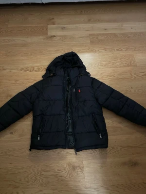 Svart pufferjacka från Polo Ralph Lauren - Svart pufferjacka från Polo Ralph Lauren med huva och dragkedja framtill. Jackan har två sidofickor med dragkedja och en liten röd logga på bröstet. Perfekt för kalla dagar och har en stilren look som funkar till det mesta.